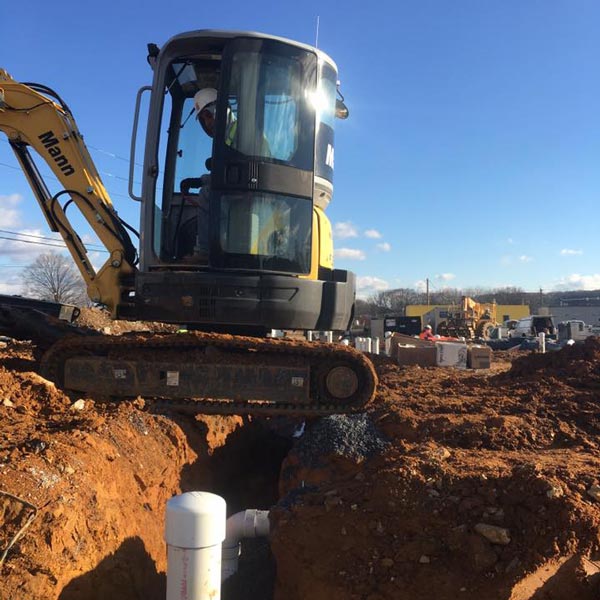 Mann Plumbing & Heating employee operating a small excavator, performing underground plumbing or heating installation.
